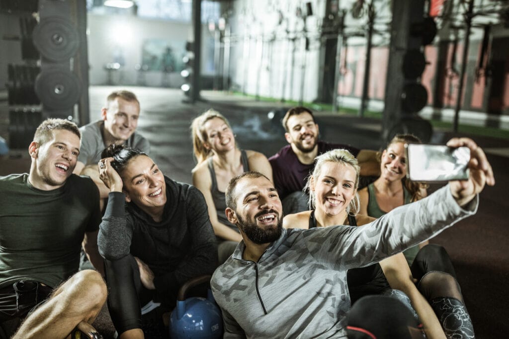 Large group of happy athletic friends taking a selfie with mobile phone on a break in a gym.